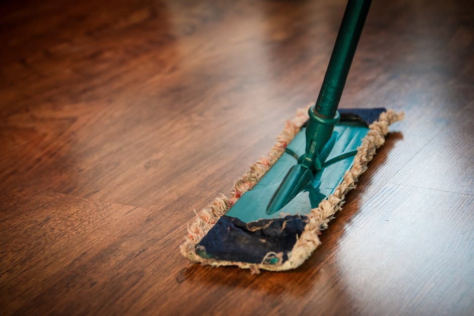 A green and black floor mop with a mop head made of worn, frayed cloth fibers is positioned on a polished wooden floor in a room. The wood surface has a warm, rich tone, and appears clean and shiny, r
