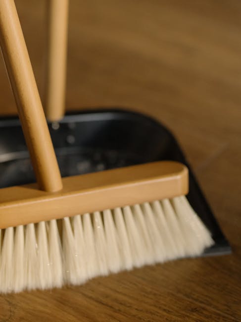 A green and black floor mop with a mop head made of worn, frayed cloth fibers is positioned on a polished wooden floor in a room. The wood surface has a warm, rich tone, and appears clean and shiny, r