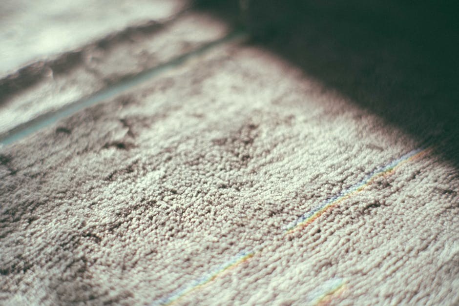 A professional cleaner wearing a white protective suit, gloves, mask, and shoe covers is engaged in deep cleaning a beige carpet in a bright, modern living room. The cleaner is using a steam or hot wa