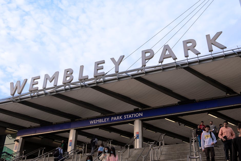 Photograph of Wembley Park station entrance under a large overhang with the bold, white 'WEMBLEY PARK' sign on top. Below, a blue sign reads 'WEMBLEY PARK STATION'. Several commuters are descending th